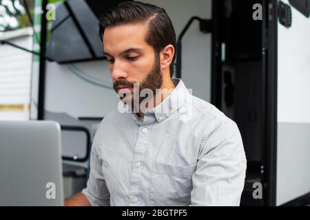A man wearing a long sleeve grey button down business shirt with brown slicked comb over hair working on laptop on a picnic table remotely near his ca Stock Photo
