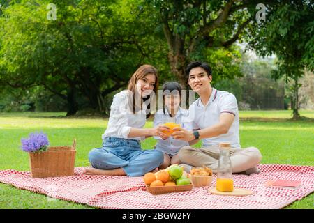 Asian teen family happy holiday picnic moment in the park with father, mother and daughter holding orange juice glass and smile to happy spend vacatio Stock Photo