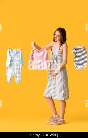 Beautiful pregnant woman hanging baby clothes on rope against color background Stock Photo