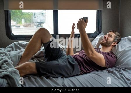 A bearded brown haired hypster man wearing a red t-shirt lays down laughing relaxing on his bed in his camper talking to someone on his smartphone on Stock Photo