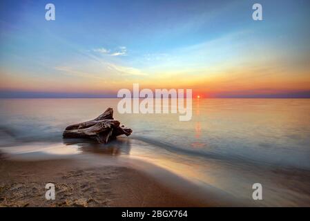 The Sunset Reflection over the Lake with the waves Moving Towards a Piece of Driftwood on the Beach Stock Photo