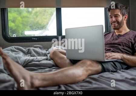 A bearded brown haired hypster man wearing a red t-shirt lays down relaxing on his bed in his camper typing and communicating to someone on his cmoput Stock Photo