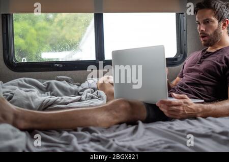 A bearded brown haired hypster man wearing a red t-shirt lays down relaxing on his bed in his camper typing and communicating to someone on his cmoput Stock Photo
