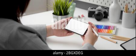 Cropped shot of professional designer using horizontal mock-up  smartphone while sitting at minimal workspace with designer supplies Stock Photo