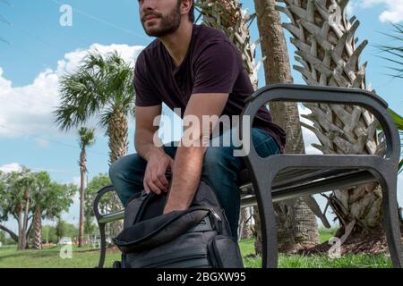 A man wearing a red shirt and blue jeans stretches his arm out reaching down into a black backpack searching on a bright sunny day on a park bench in Stock Photo