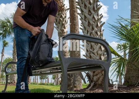 A man wearing a red shirt and blue jeans stretches his arm out reaching down into a black backpack searching on a bright sunny day on a park bench in Stock Photo