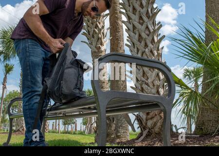 A man wearing a red shirt and blue jeans stretches his arm out reaching down into a black backpack searching on a bright sunny day on a park bench in Stock Photo