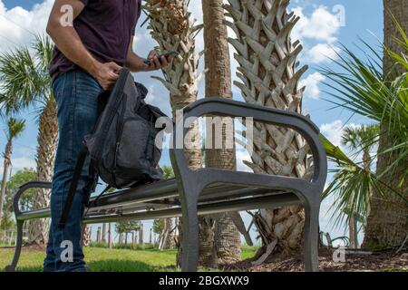 A man wearing a red shirt and blue jeans stretches his arm out reaching down into a black backpack searching on a bright sunny day on a park bench tex Stock Photo