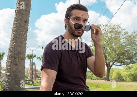 A man wearing a red shirt and blue jeans sits down slightly taking his silver aviators off looking at something passing by in awe capturing all his at Stock Photo