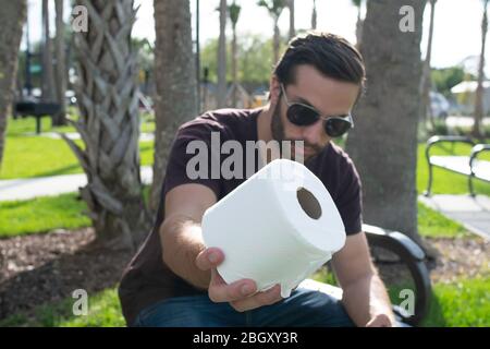 A man with a red shirt and blue jeans relaxes on a park bench Holding up precious roll of toilet paper like there's not enough to go around during a s Stock Photo