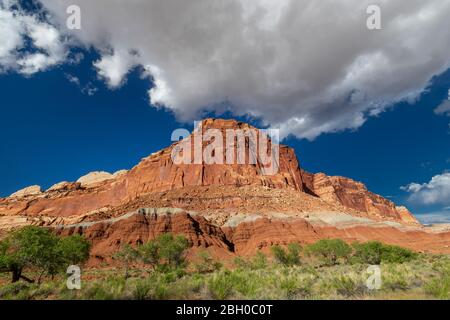 Landscape shot of a large red sandstone formation sloping down on both sides, with a similarly shaped puffy cloud just above it, against a solid blue Stock Photo