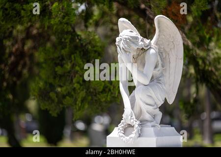 Close up of the white marble statue of a kneeling winged angel holding a laurel crown and crying, against a green bokeh background Stock Photo