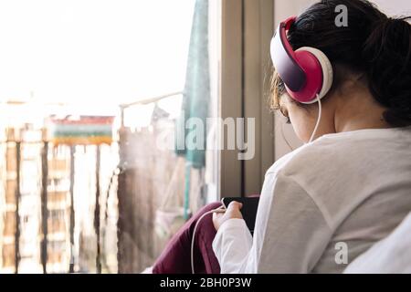 little girl sitting in front of her house balcony with headset watching videos on the phone, home entertainment for children concept, copy space for t