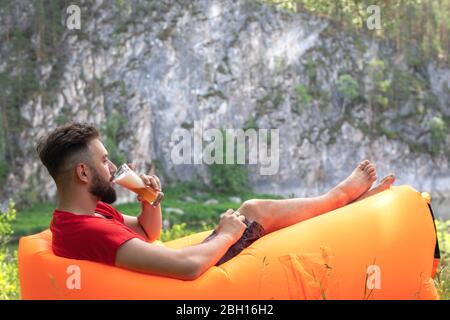 Bearded man sitting with glass of beer in hand enjoying sunny day. Young male camper in nature drinking beer and enjoying weather. Summer holidays con Stock Photo