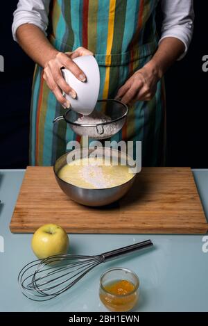 Working in the kitchen. Going through a flour sieve with a strainer. Cooking apple pie. Colorful apron. Steel bowl. White glass kitchen table. Dark bl Stock Photo