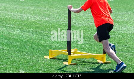 A teenage boy at a running camp is pushind a yellow weight sled on a green turf field. Stock Photo