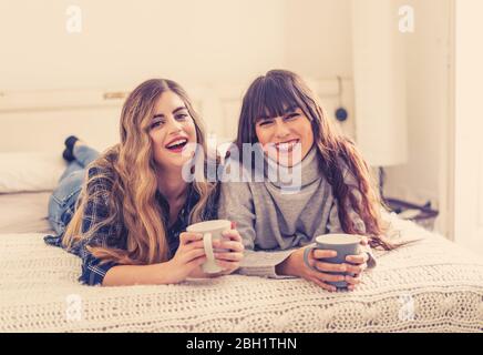 COVID-19 lockdown. Confident and optimistic women best girlfriends talking and laughing in bed with hot drink staying together at home in isolation. S Stock Photo