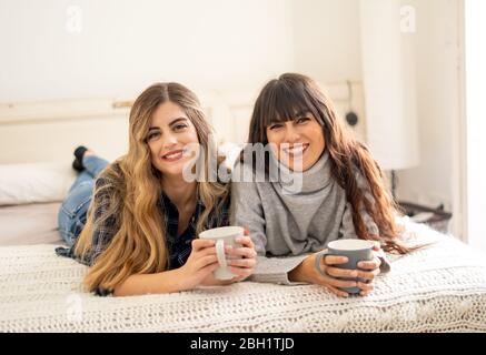 COVID-19 lockdown. Confident and optimistic women best girlfriends talking and laughing in bed with hot drink staying together at home in isolation. S Stock Photo