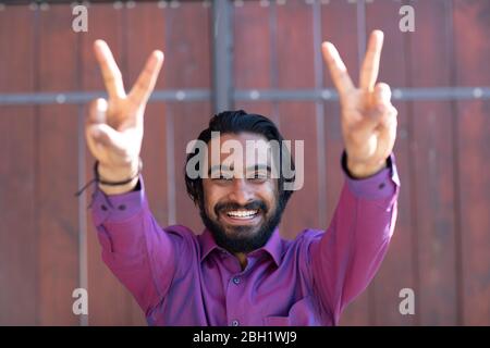 Young smiling man making peace sign in front of a gate Stock Photo