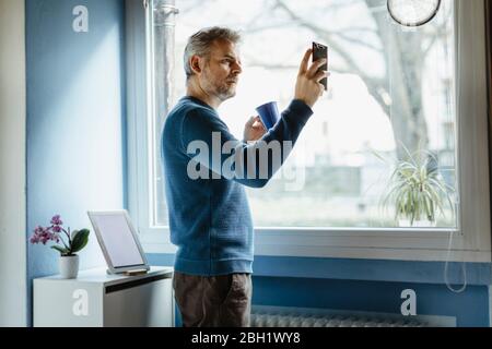 Mature man with coffee mug standing in living room taking selfie with smartphone Stock Photo