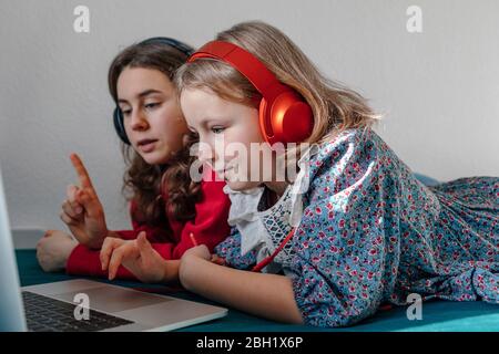 Two sisters with headphones lying together on bed looking at laptop Stock Photo