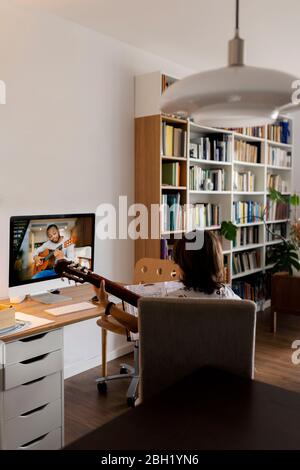 Back view of boy sitting on arm chair at home using online tutorial for learning to play guitar Stock Photo