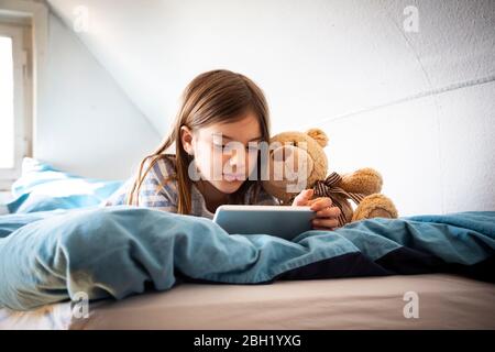 Portrait of girl lying on bed with teddy bear using digital tablet Stock Photo