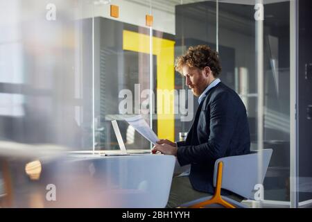 Businessman sitting at desk in office with laptop reading papers Stock Photo