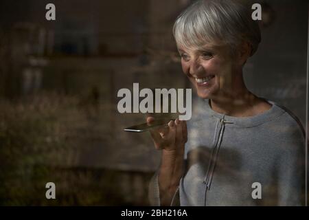 Portrait of smiling senior woman behind windowpane using smartphone Stock Photo