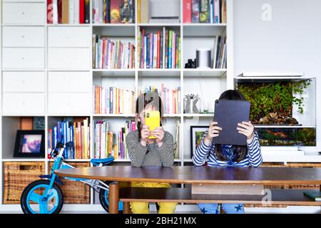 Two sisters sitting side by side at table in the living room using electronic devices Stock Photo
