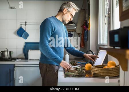 Mature man preparing artichokes in his kitchen using digital tablet Stock Photo