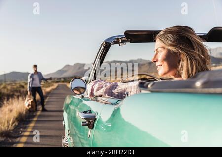 Woman in convertible car on a road trip with man hitchhiking at the roadside Stock Photo