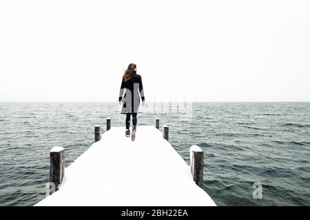 Back view of young woman walking on snow-covered jetty at Lake Starnberg, Germany Stock Photo