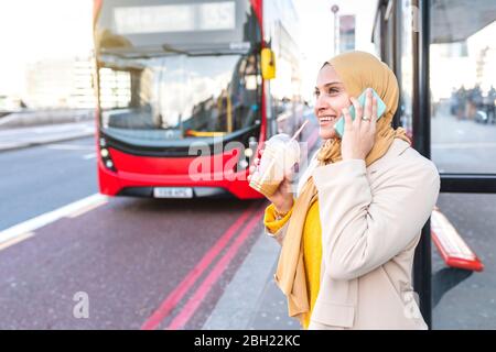Portrait of smiling young woman on the phone waiting at bus stop Stock Photo