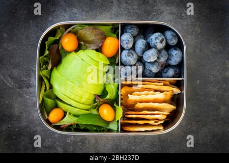 Lunch box with sliced avocado, yellow tomatoes, crackers, blueberries and green salad Stock Photo