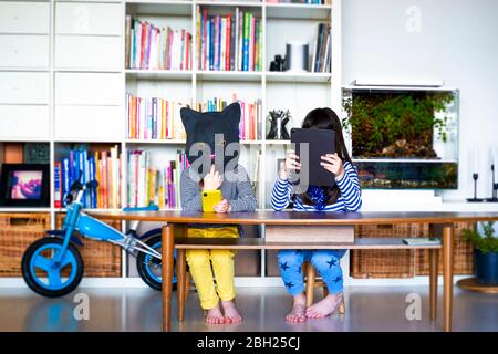 Two girls sitting at table, hiding behind digital tablet and cat mask Stock Photo