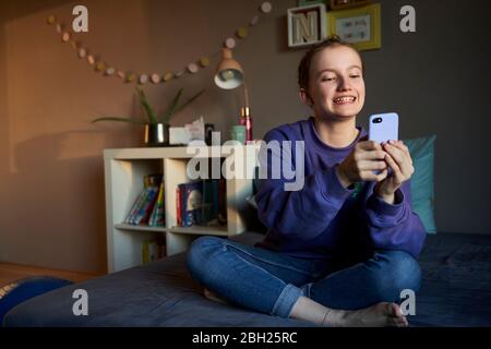 Portrait of grinning girl sitting on bed taking selfie with smartphone Stock Photo