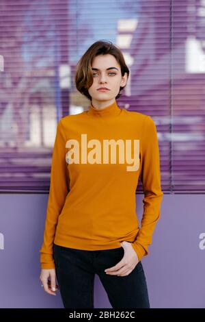 Portrait of young woman wearing orange turtleneck pullover, purple glass wall in the background Stock Photo