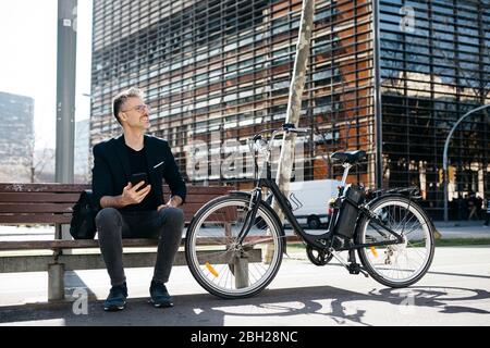 Gray-haired businessman sitting on a bench next to electric bicycle in the city Stock Photo