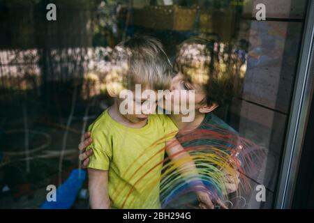Mother kissing her little son behind window with drawn rainbow Stock Photo
