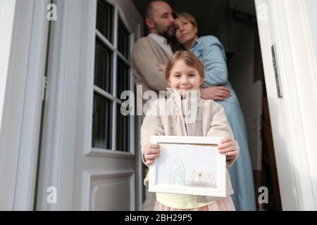 Happy little girl with drawing of her house and parents hugging behind her Stock Photo