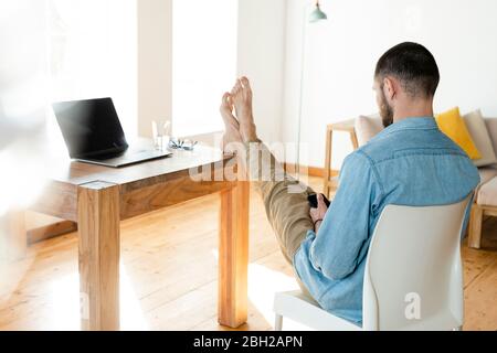 Young man at home typing on his smartphone in modern living room with his feet on the table Stock Photo