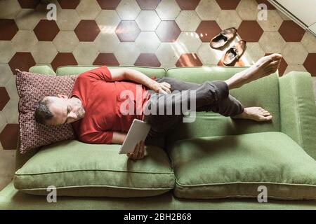 Mature man lying on sofa in living room and using tablet Stock Photo