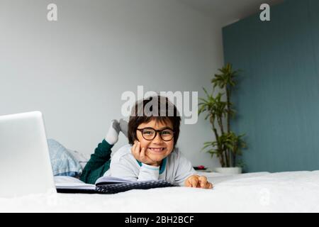 Portrait of relaxed little boy lying on bed with laptop doing homework Stock Photo