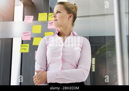 Young businesswoman, standing in front of adhesive notes, with arms crossed Stock Photo