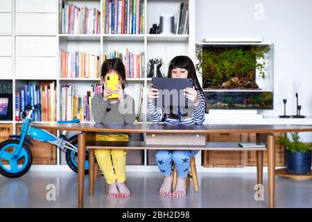Two sisters sitting side by side at table in the living room using electronic devices Stock Photo