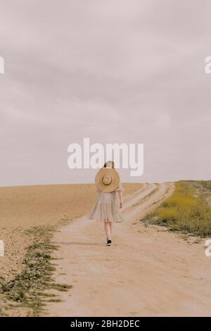 Woman with straw hat and vintage dress walking on a remote field road in the countryside Stock Photo