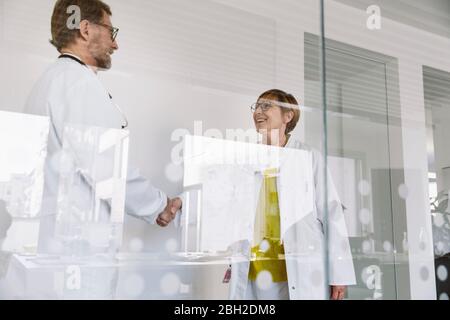 Two smiling doctors shaking hands behind glass wall Stock Photo