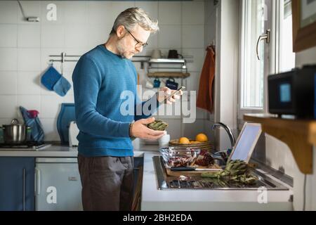 Mature man preparing artichoke in his kitchen looking at smartphone Stock Photo