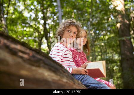 Children with book sitting on log in forest Stock Photo